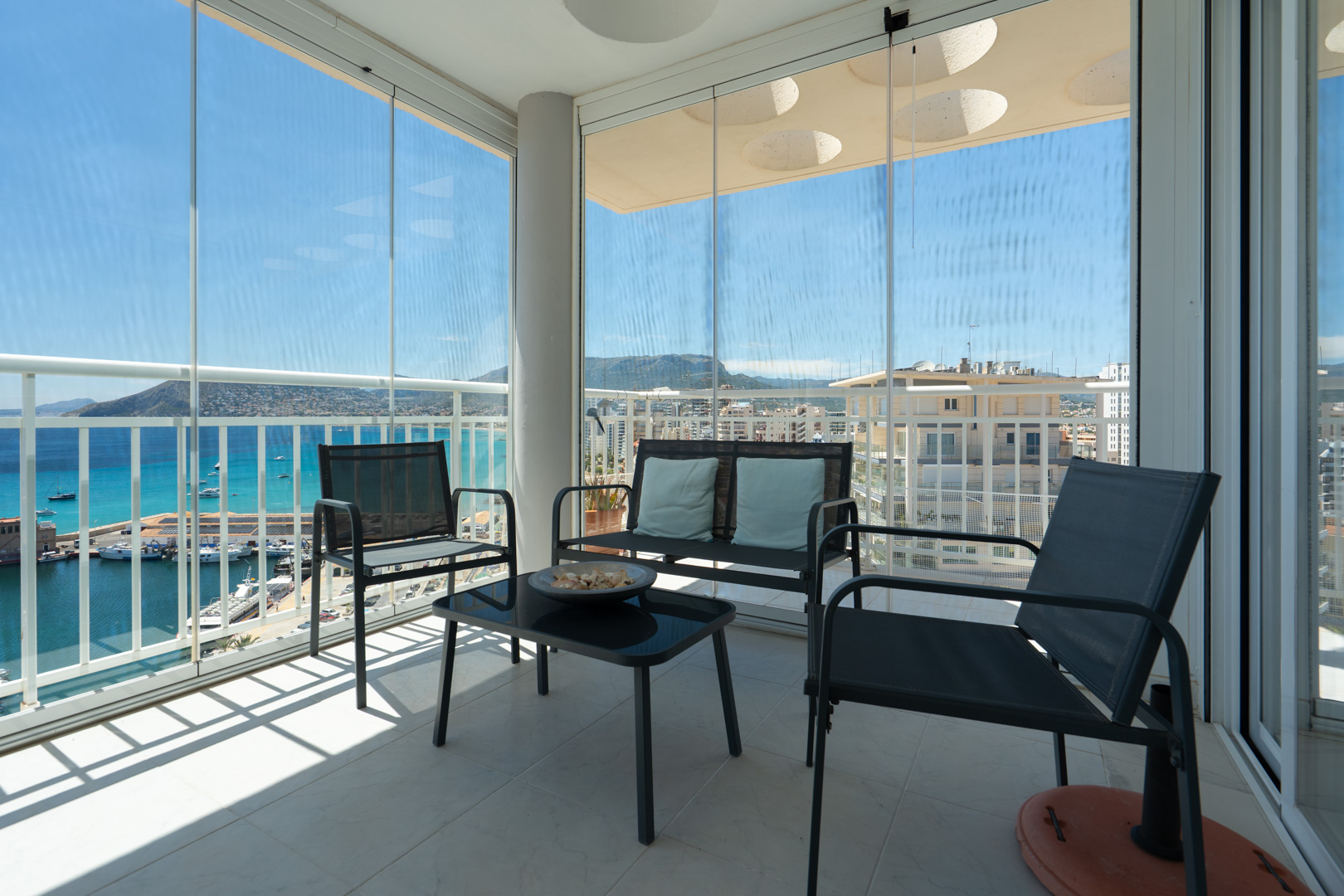 Terrasse couverte avec rideaux de verre coulissants, mobilier d'extérieur et vues panoramiques sur le port de Calpe, la mer et les montagnes. Le sol est carrelé.