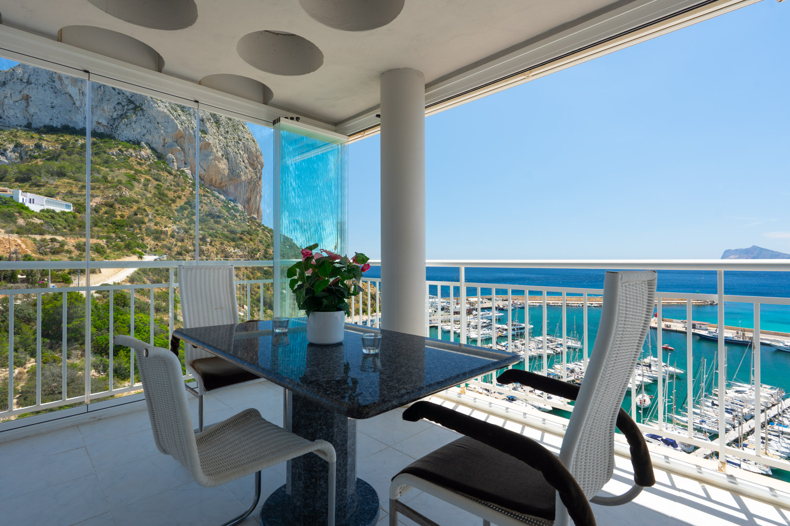 Terrasse couverte avec table en granit et chaises modernes, dotée de rideaux de verre, offrant des vues panoramiques sur le Peñón de Ifach et le port de Calpe.