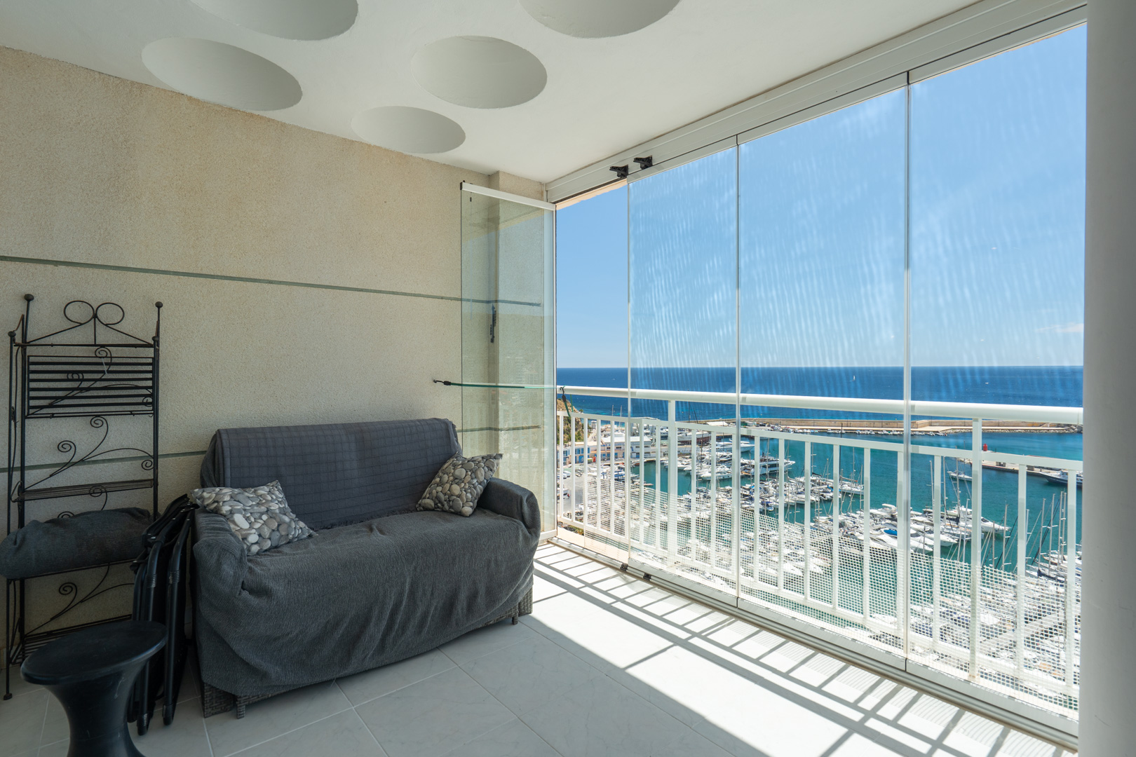 Terrasse vitrée offrant une vue imprenable sur le port de plaisance et la mer, aménagée avec un canapé et une étagère en fer forgé, sous un plafond à motifs circulaires.
