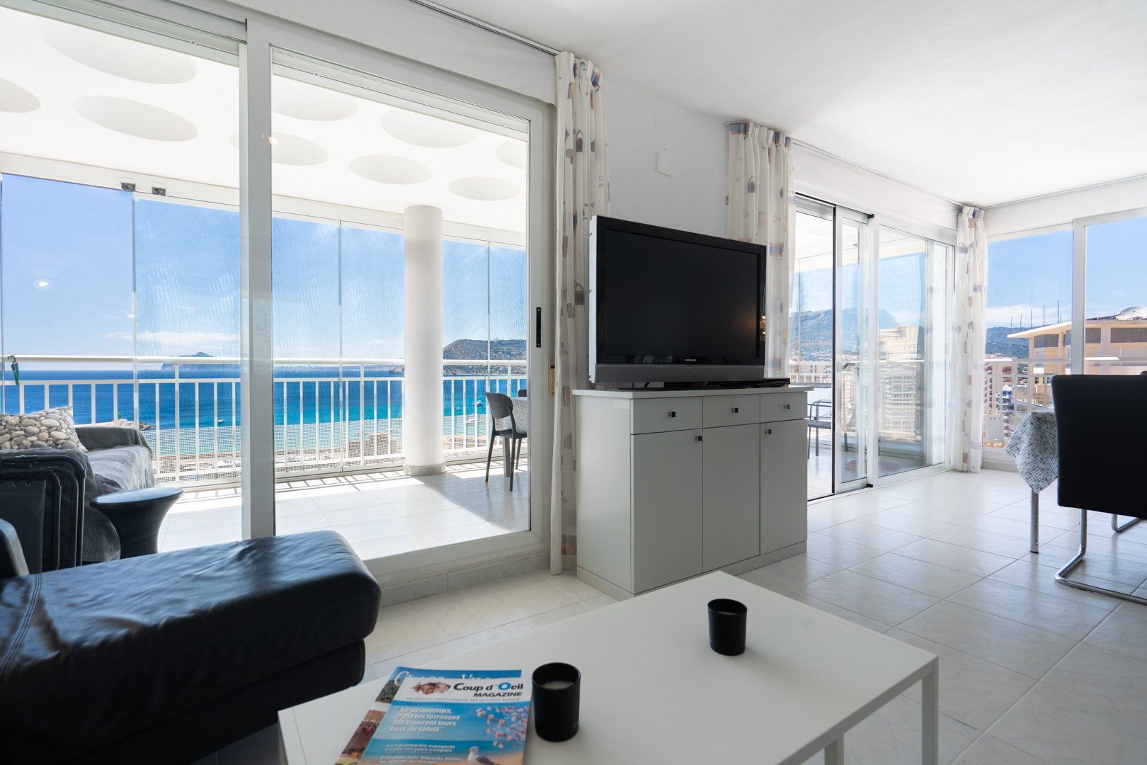 Vaste salon carrelé avec meuble TV et canapé, s'ouvrant sur une terrasse vitrée offrant une vue mer panoramique et un plafond à motifs circulaires.