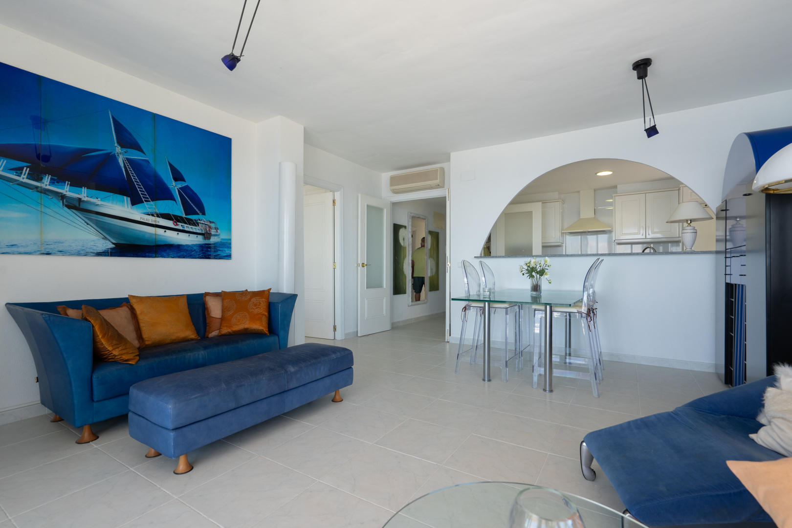 Vaste salon-salle à manger avec canapé bleu, table en verre et chaises transparentes, agrémenté d'une grande toile de voilier. Une arche ouvre sur la cuisine semi-ouverte moderne.