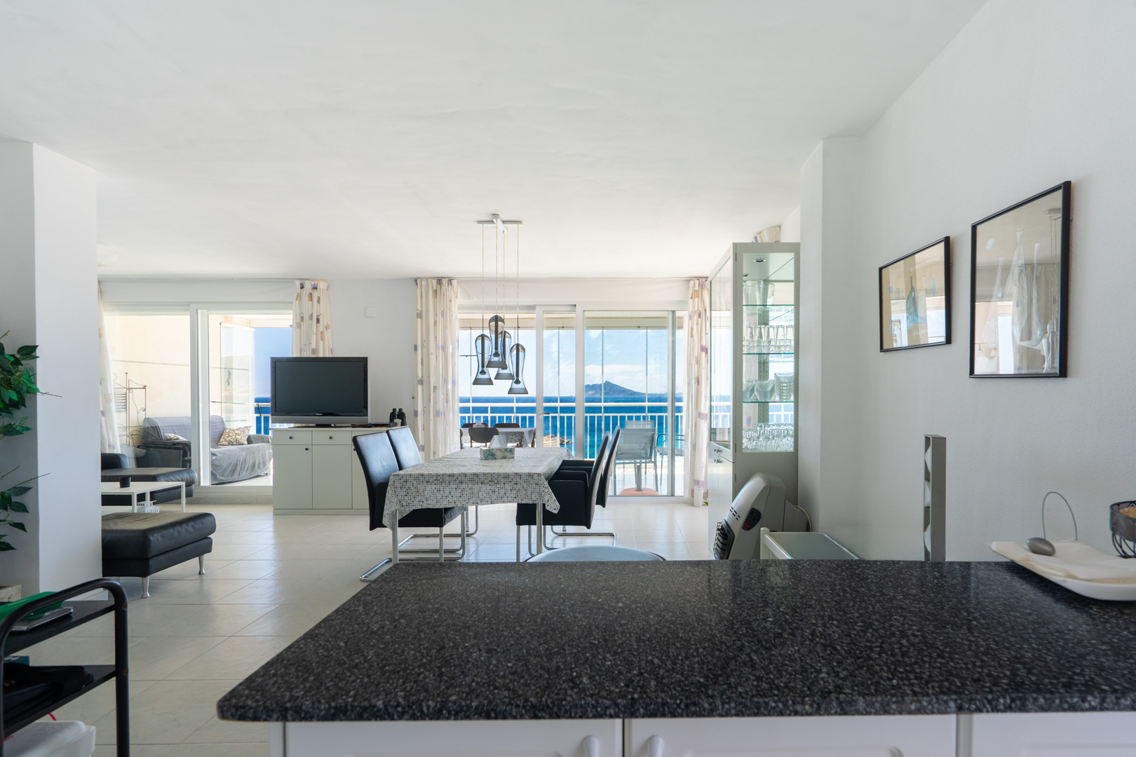 Vaste salon-salle à manger ouvert, avec suspension moderne au-dessus de la table, sol carrelé clair et accès direct à une terrasse offrant une vue imprenable sur la mer par des portes coulissantes.