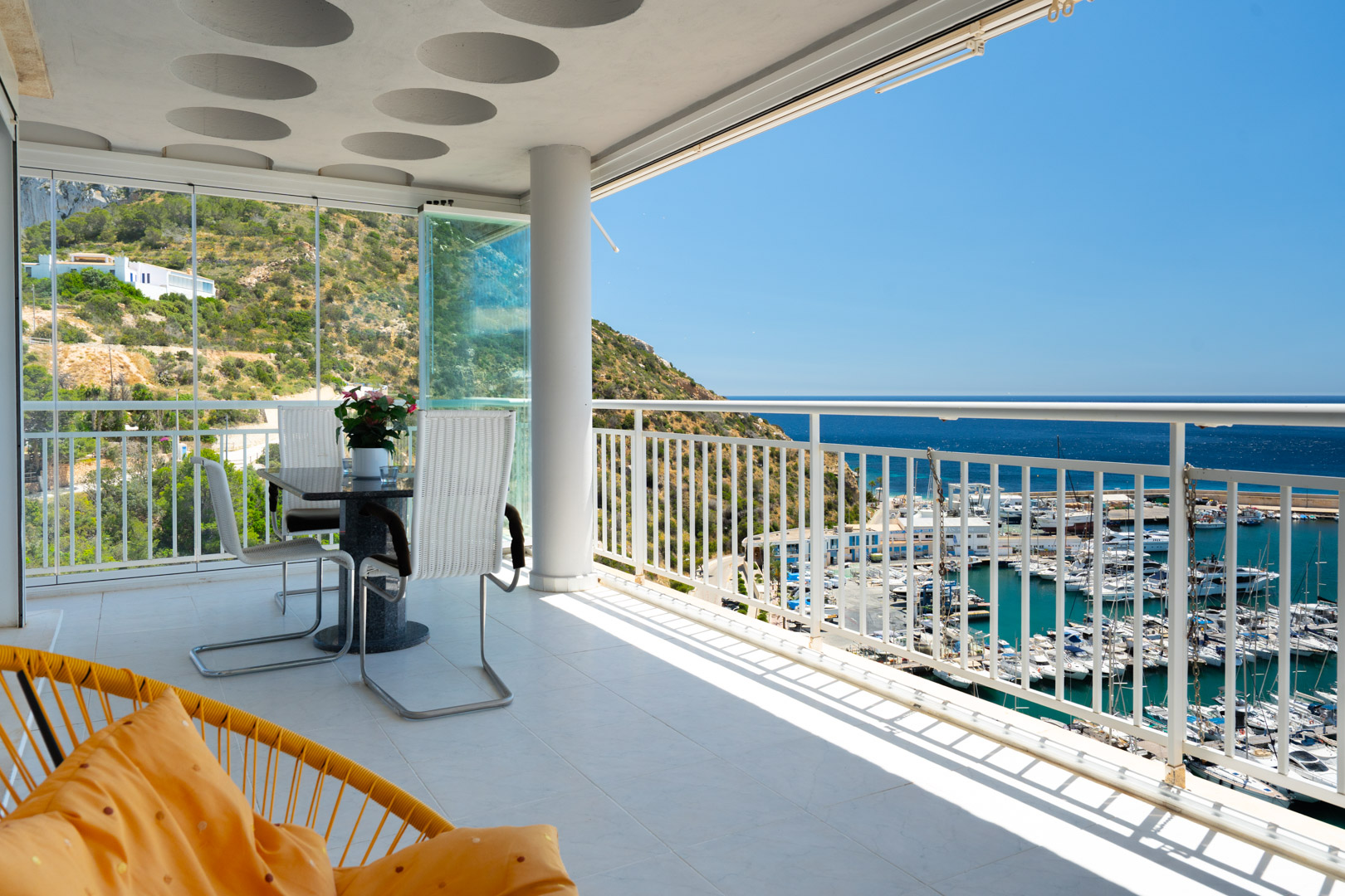 Vaste terrasse carrelée avec table à manger et rideaux de verre, offrant une vue panoramique directe sur le port de plaisance de Calpe, la mer Méditerranée et les collines verdoyantes.