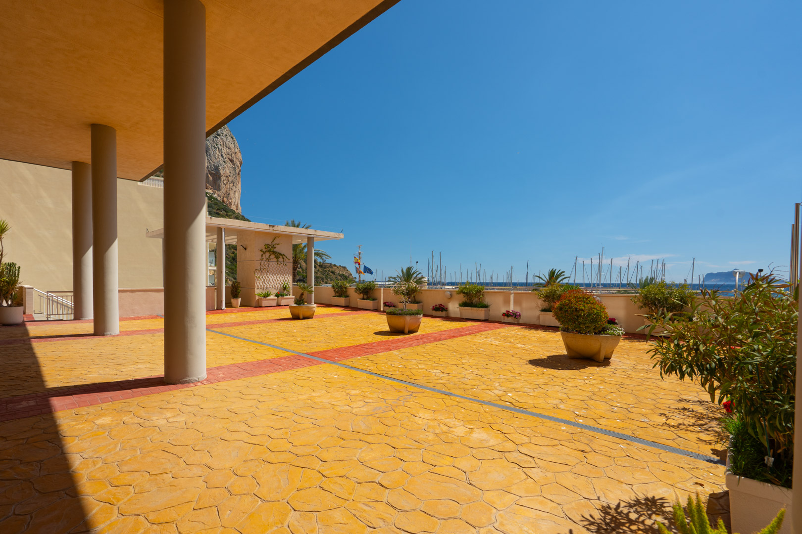 Vaste terrasse commune avec pavé texturé jaune distinctif et accents rouges, offrant une vue directe sur la marina avec ses nombreux mâts de bateaux et l'emblématique Peñón de Ifach sous un ciel clair.