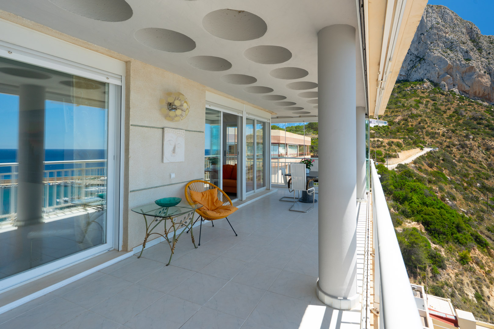 Vaste terrasse couverte offrant des vues panoramiques sur la mer étincelante et l'imposant Peñón de Ifach. Elle présente un plafond distinctif avec des cercles encastrés et un sol carrelé.