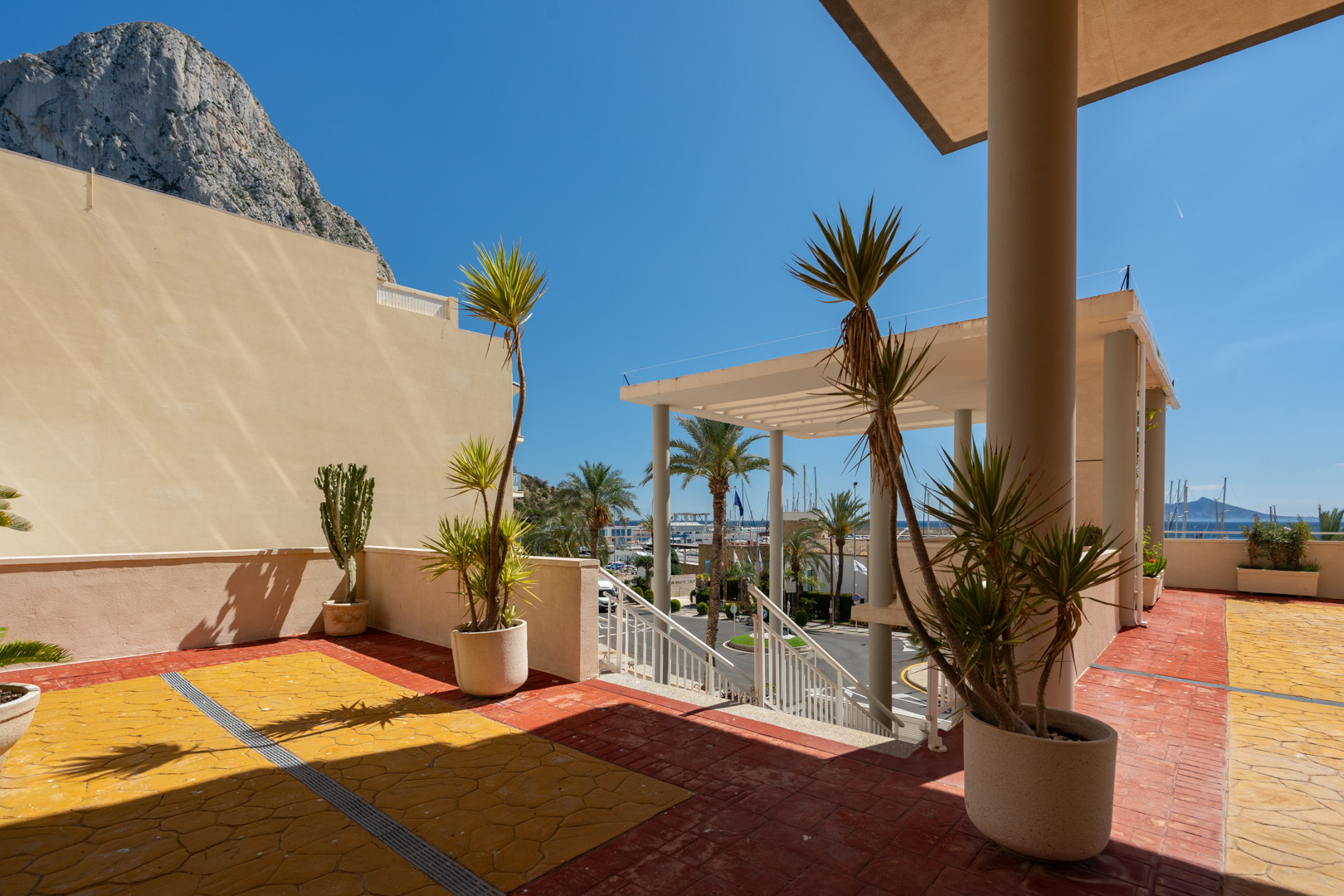 Vaste terrasse extérieure avec carrelage rouge et jaune, plantes méditerranéennes, offrant une vue directe sur le port de Calpe et le Peñón de Ifach sous un ciel bleu.