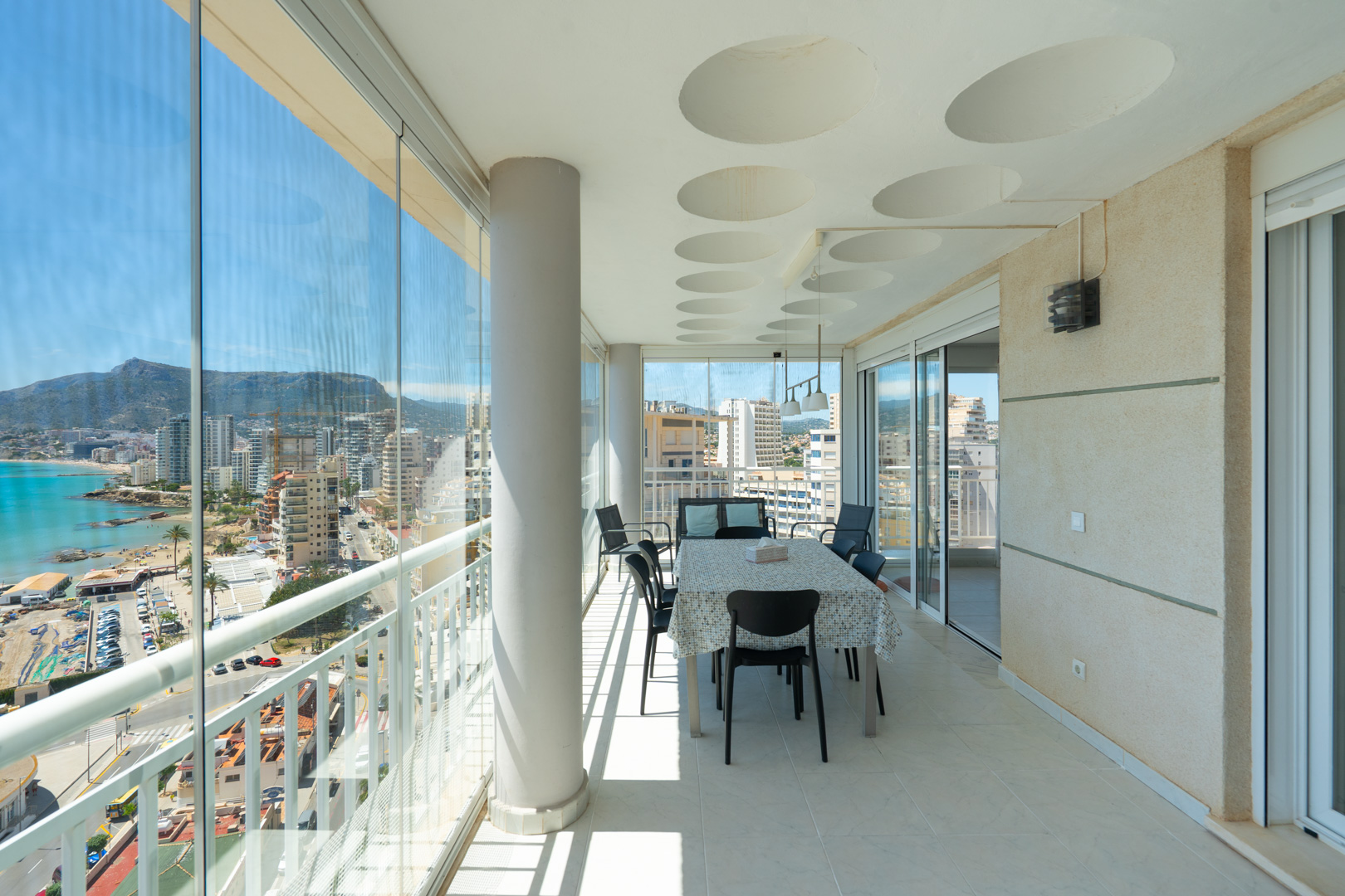 Vaste terrasse vitrée avec salle à manger, offrant des vues panoramiques sur la mer, le port et les montagnes de Calpe. Elle dispose de rideaux de verre, d'un plafond à motifs circulaires et d'un sol carrelé.