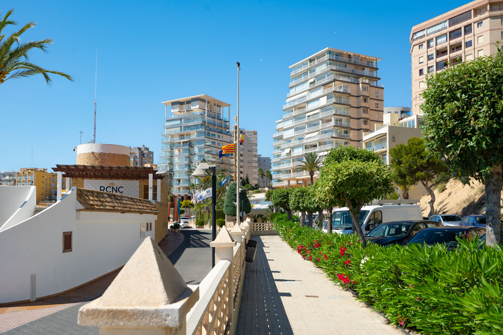 Vue du quartier animé du port de Calpe, présentant des immeubles résidentiels modernes, une promenade pavée bordée de buissons verts et de fleurs rouges, et le bâtiment RCNC, sous un ciel bleu.