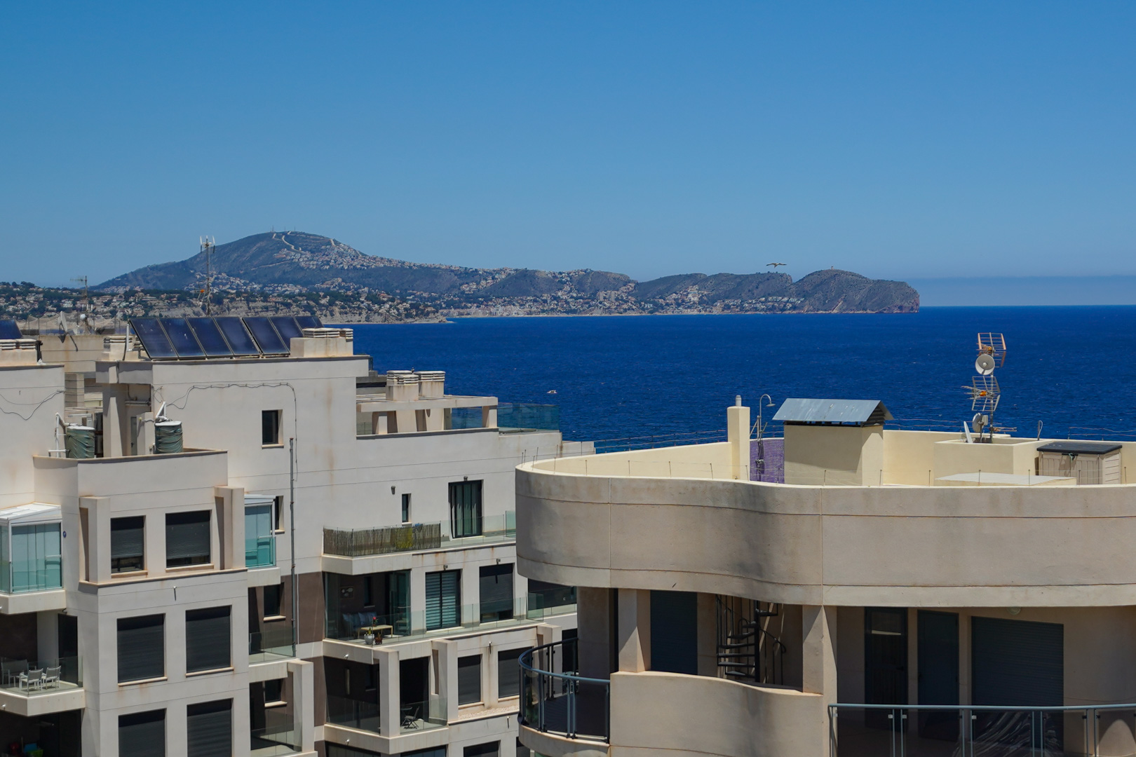 Vue élevée sur les toits avec panneaux solaires, la mer d'un bleu profond et une chaîne de montagnes côtière lointaine sous un ciel clair.