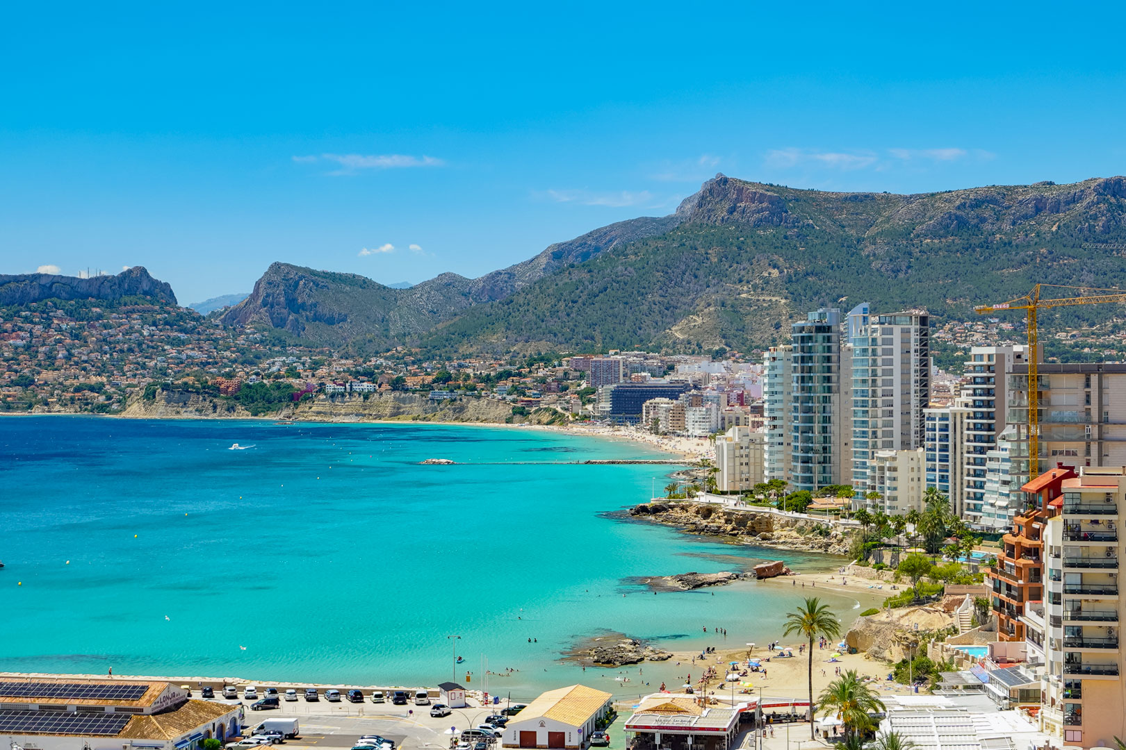 Vue panoramique de la baie de Calpe avec ses eaux turquoise, plages de sable, bâtiments côtiers et montagnes lointaines sous un ciel bleu éclatant.