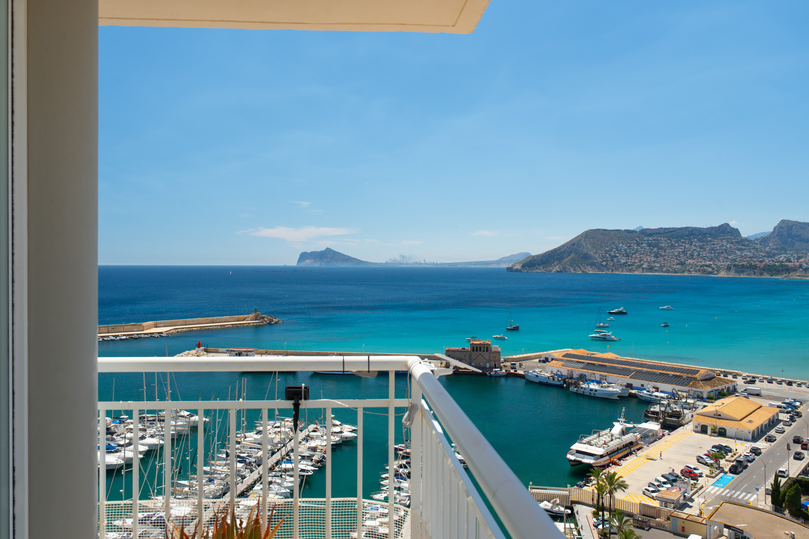 Vue panoramique depuis le balcon sur la marina de Calpe, ses yachts, la baie aux eaux turquoise et les montagnes côtières lointaines sous un ciel clair.