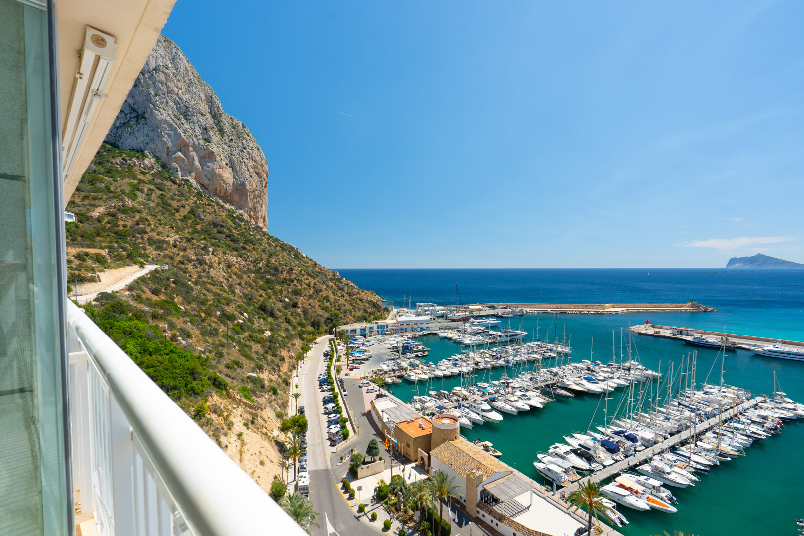 Vue panoramique depuis un balcon, dévoilant le port de plaisance de Calpe rempli de bateaux, la mer turquoise éclatante et l'imposant Peñón de Ifach sous un ciel azur.