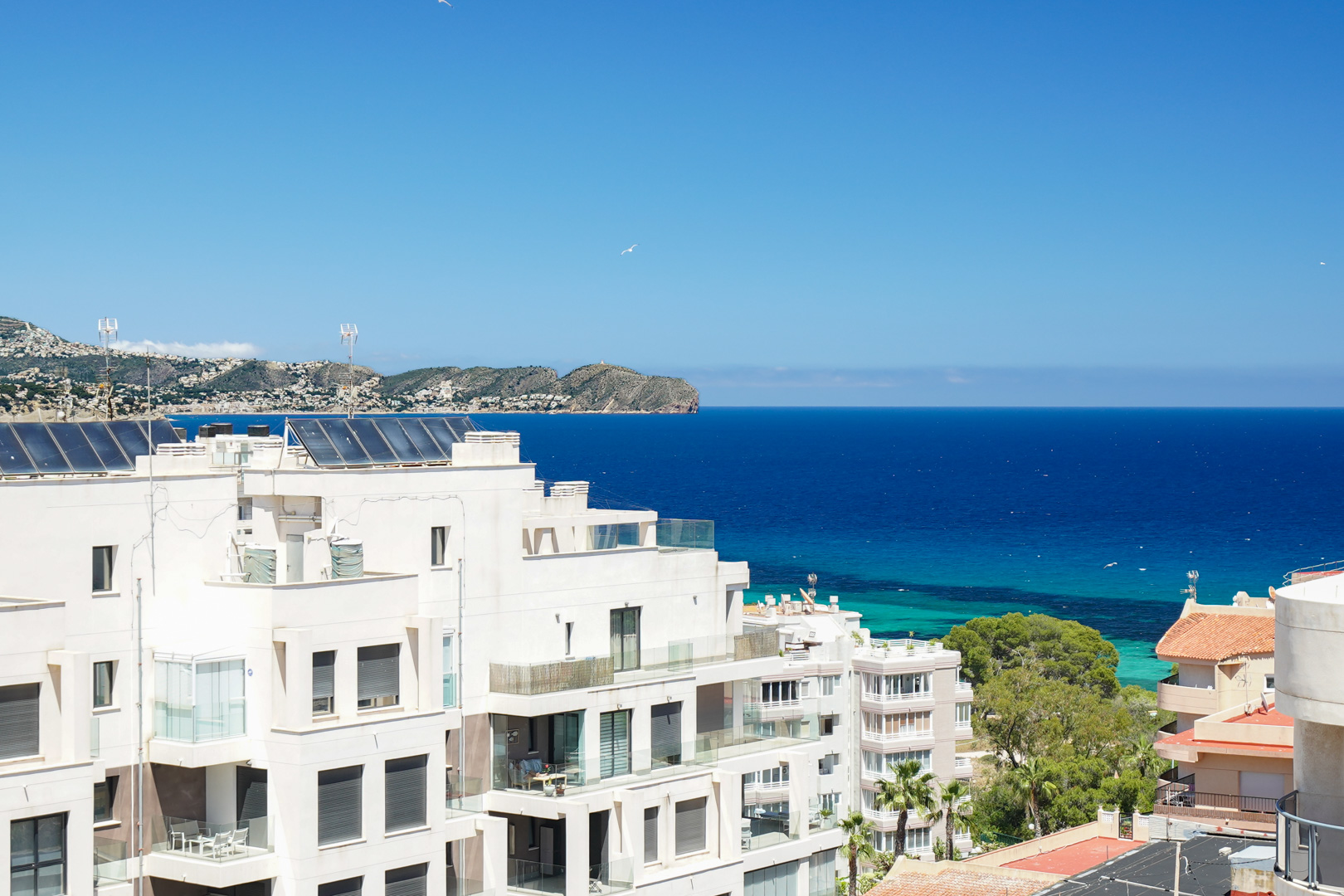 Vue panoramique sur des bâtiments résidentiels blancs modernes avec balcons vitrés, panneaux solaires en toiture et la mer Méditerranée d'un bleu profond jusqu'à la côte lointaine.