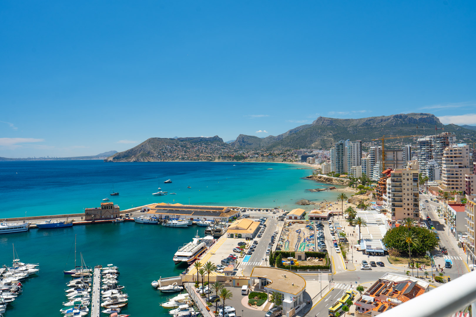 Vue panoramique sur la baie de Calpe, le port de plaisance animé, les plages de sable fin et le littoral urbain, avec les montagnes en toile de fond sous un ciel azur.