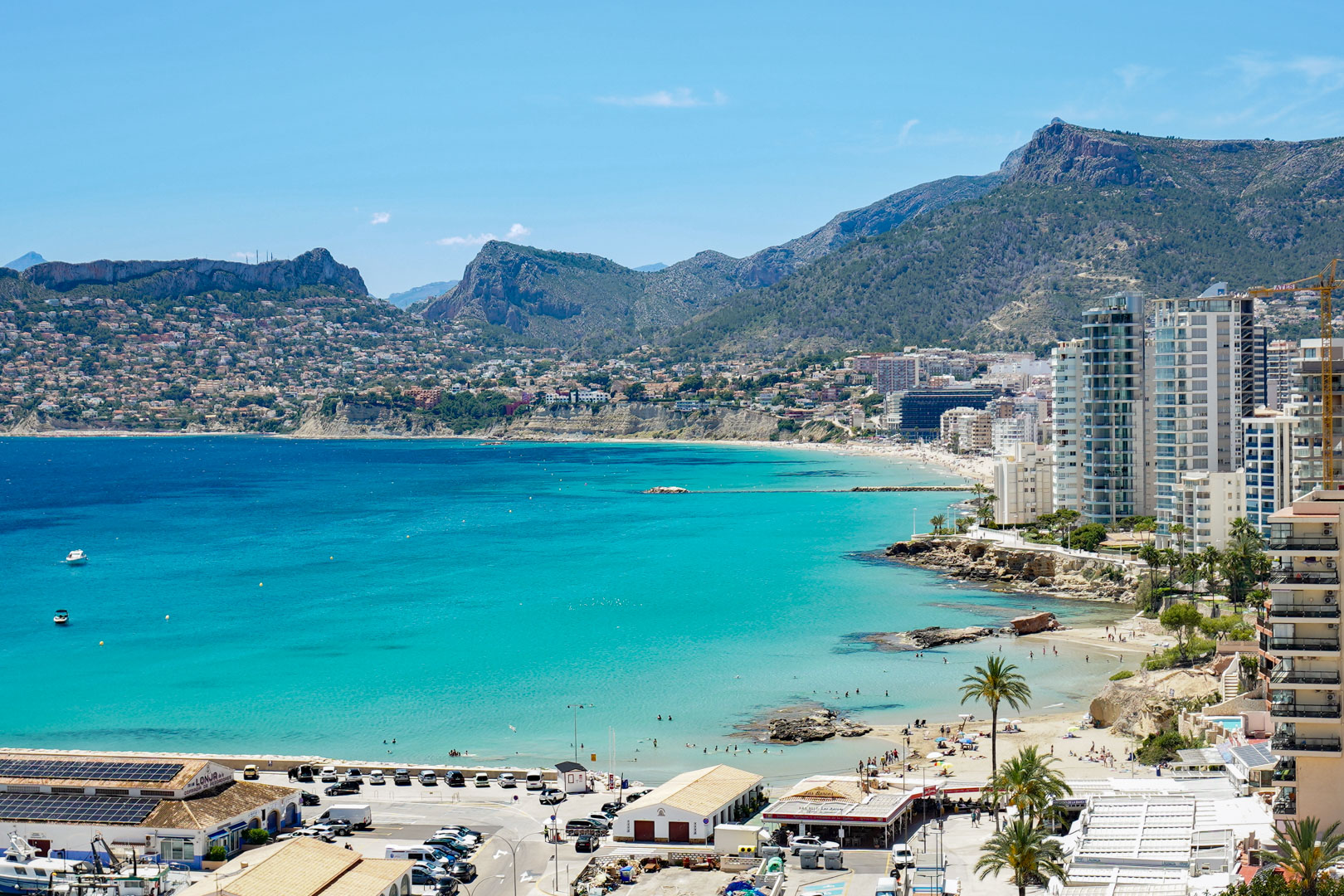 Vue panoramique sur la baie turquoise de Calpe, présentant des plages de sable, des immeubles côtiers et des montagnes lointaines sous un ciel clair.