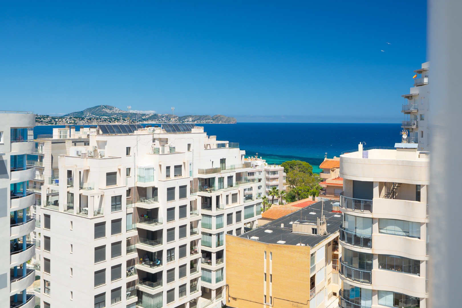 Vues panoramiques sur la mer Méditerranée et la côte depuis un étage élevé, avec des bâtiments résidentiels au premier plan et des montagnes lointaines sous un ciel bleu clair.