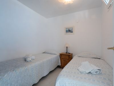 Chambre lumineuse avec deux lits simples aux parures claires, une table de chevet en bois avec lampe, un tableau décoratif et une fenêtre en hauteur.