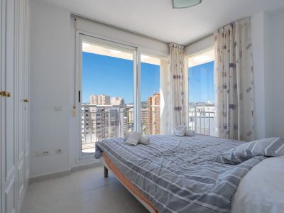Chambre lumineuse avec lit double, armoire intégrée et portes coulissantes vers une terrasse. Vues urbaines dégagées sous un ciel bleu.