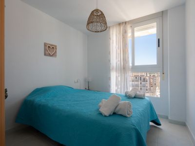 Chambre lumineuse avec lit double en parure turquoise, suspension en fibres naturelles et fenêtre offrant une vue urbaine sur les bâtiments environnants.