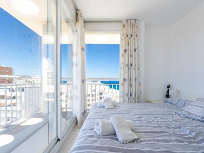 Chambre lumineuse avec lit double et linge de lit rayé, offrant un accès direct à une terrasse avec vue panoramique sur la mer et le ciel.