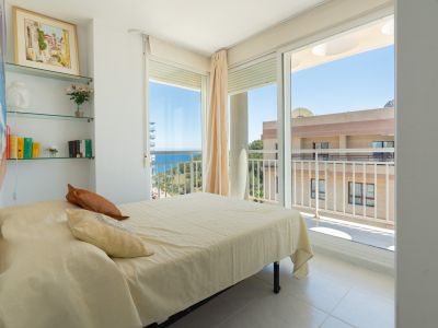 Chambre lumineuse avec lit double, étagères en verre décoratives et accès direct à une terrasse offrant une vue partielle sur la mer.