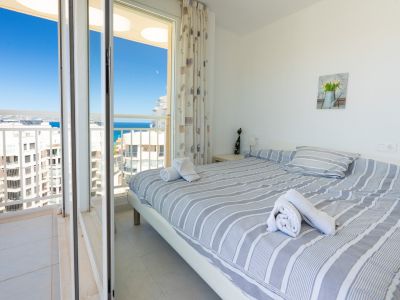 Chambre lumineuse avec lit double, linge de lit rayé, lampe de chevet et accès direct à un balcon privé offrant vue sur mer et ville.