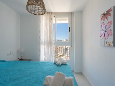 Chambre lumineuse avec un lit au couvre-lit bleu matelassé et serviettes blanches. Fenêtre offrant des vues urbaines et montagneuses, suspension en osier et tableau floral.