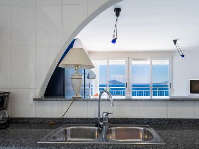 Coin cuisine avec évier double en inox et plan de travail foncé, s'ouvrant sur un salon lumineux et une terrasse offrant une vue imprenable sur la mer et la côte.