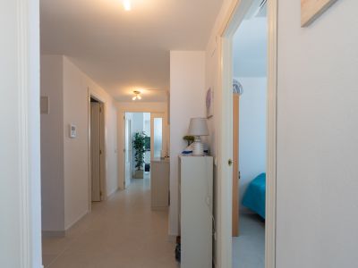 Couloir lumineux avec carrelage clair, portes blanches et une console avec lampe. Aperçu d'une chambre avec lit bleu et d'une autre pièce avec plante.