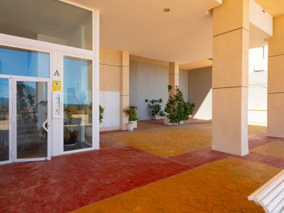 Entrée principale avec portes vitrées à cadre blanc, sol pavé bicolore rouge et orange, et piliers de soutien beiges. Des plantes en pot agrémentent cet espace extérieur couvert.