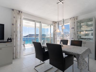 Salle à manger lumineuse avec table et chaises modernes, ouvrant sur une terrasse vitrée offrant des vues panoramiques sur la mer et le Peñón de Ifach. Vitrine à vaisselle.