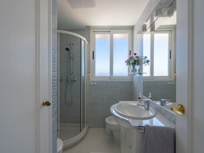 Salle de bain avec cabine de douche d'angle, meuble-lavabo blanc en marbre et carreaux de mosaïque bleu clair. Fenêtre et miroir éclairé par des spots.