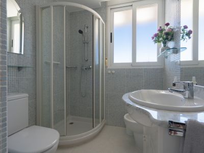Salle de bain avec douche d'angle, toilettes, bidet et lavabo sur meuble effet marbre. Les murs sont recouverts de mosaïque bleu clair, éclairée par des fenêtres.