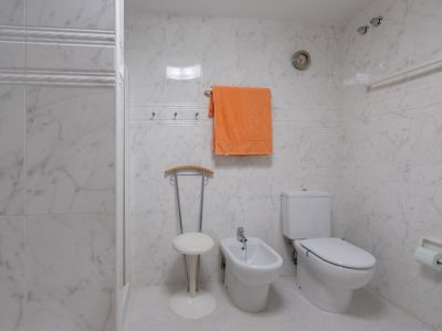 Salle de bain carrelée effet marbre blanc, comprenant des toilettes, un bidet et un espace douche fonctionnel avec barre d'appui. Un horloge murale et des patères sont également présents.
