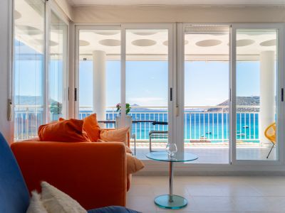 Salon lumineux avec canapé orange et table en verre, s'ouvrant sur une terrasse par de grandes portes coulissantes offrant une vue directe sur la mer, les bateaux et montagnes.