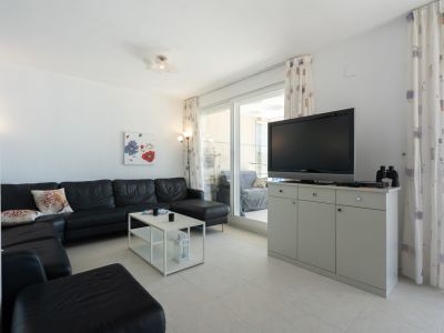 Salon spacieux avec canapé d'angle en cuir noir, table basse blanche et carrelage au sol. Des portes coulissantes en verre mènent à une terrasse, complété par un meuble TV et des rideaux à motifs.