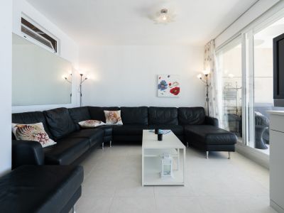 Salon spacieux avec un grand canapé d'angle noir et coussins décoratifs, sol carrelé blanc et table basse moderne. Les portes coulissantes vitrées s'ouvrent sur une terrasse.