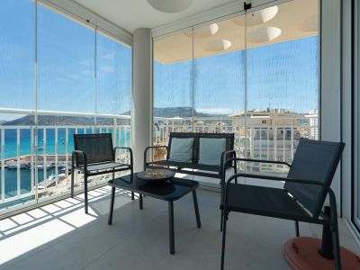 Terrasse couverte avec rideaux de verre coulissants, mobilier d'extérieur et vues panoramiques sur le port de Calpe, la mer et les montagnes. Le sol est carrelé.