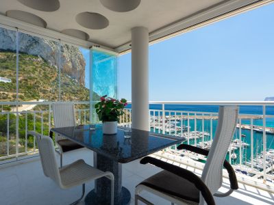Terrasse couverte avec table en granit et chaises modernes, dotée de rideaux de verre, offrant des vues panoramiques sur le Peñón de Ifach et le port de Calpe.