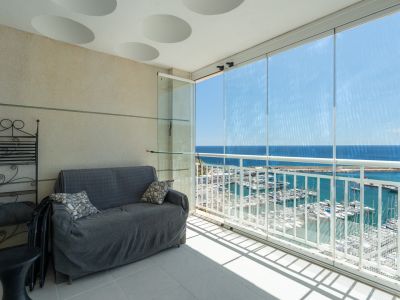 Terrasse vitrée offrant une vue imprenable sur le port de plaisance et la mer, aménagée avec un canapé et une étagère en fer forgé, sous un plafond à motifs circulaires.