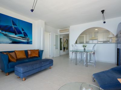 Vaste salon-salle à manger avec canapé bleu, table en verre et chaises transparentes, agrémenté d'une grande toile de voilier. Une arche ouvre sur la cuisine semi-ouverte moderne.