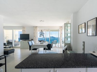 Vaste salon-salle à manger ouvert, avec suspension moderne au-dessus de la table, sol carrelé clair et accès direct à une terrasse offrant une vue imprenable sur la mer par des portes coulissantes.