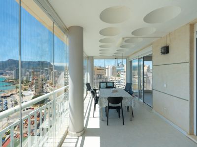 Vaste terrasse vitrée avec salle à manger, offrant des vues panoramiques sur la mer, le port et les montagnes de Calpe. Elle dispose de rideaux de verre, d'un plafond à motifs circulaires et d'un sol carrelé.