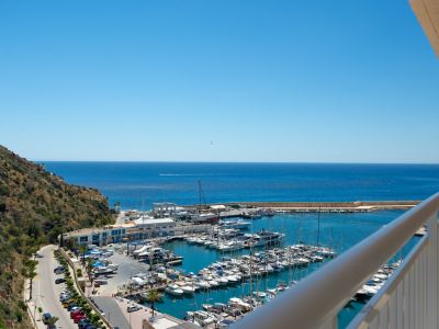 Vue panoramique depuis un balcon sur un port de plaisance animé, la mer d'un bleu profond et une colline verdoyante sous un ciel clair.