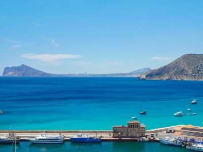 Vue panoramique du port de Calpe avec ses bateaux amarrés, les eaux turquoise et l'emblématique Peñón de Ifach en arrière-plan, bordé par la côte urbanisée.