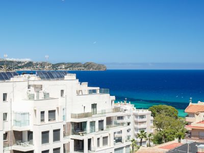 Vue panoramique sur des bâtiments résidentiels blancs modernes avec balcons vitrés, panneaux solaires en toiture et la mer Méditerranée d'un bleu profond jusqu'à la côte lointaine.