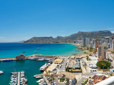 Vue panoramique sur la baie de Calpe, le port de plaisance animé, les plages de sable fin et le littoral urbain, avec les montagnes en toile de fond sous un ciel azur.