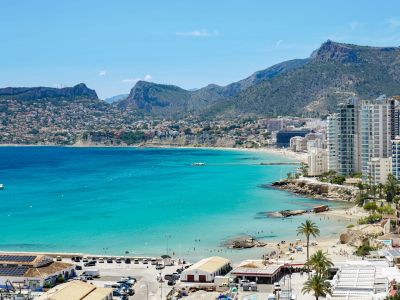 Vue panoramique sur la baie turquoise de Calpe, présentant des plages de sable, des immeubles côtiers et des montagnes lointaines sous un ciel clair.