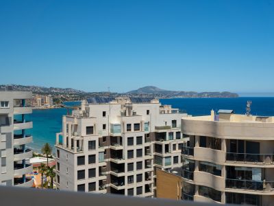 Vue panoramique sur la mer depuis une terrasse en étage élevé, présentant une architecture côtière moderne, des eaux turquoise et des montagnes lointaines sous un ciel clair.