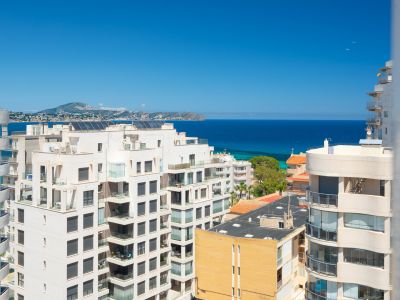 Vues panoramiques sur la mer Méditerranée et la côte depuis un étage élevé, avec des bâtiments résidentiels au premier plan et des montagnes lointaines sous un ciel bleu clair.